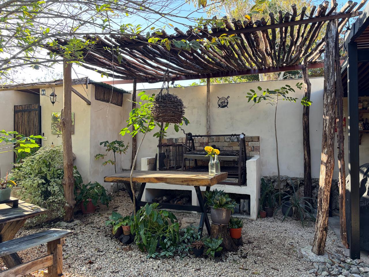 Patio exterior con pérgola de madera, área de barbacoa y mesa rústica con plantas.