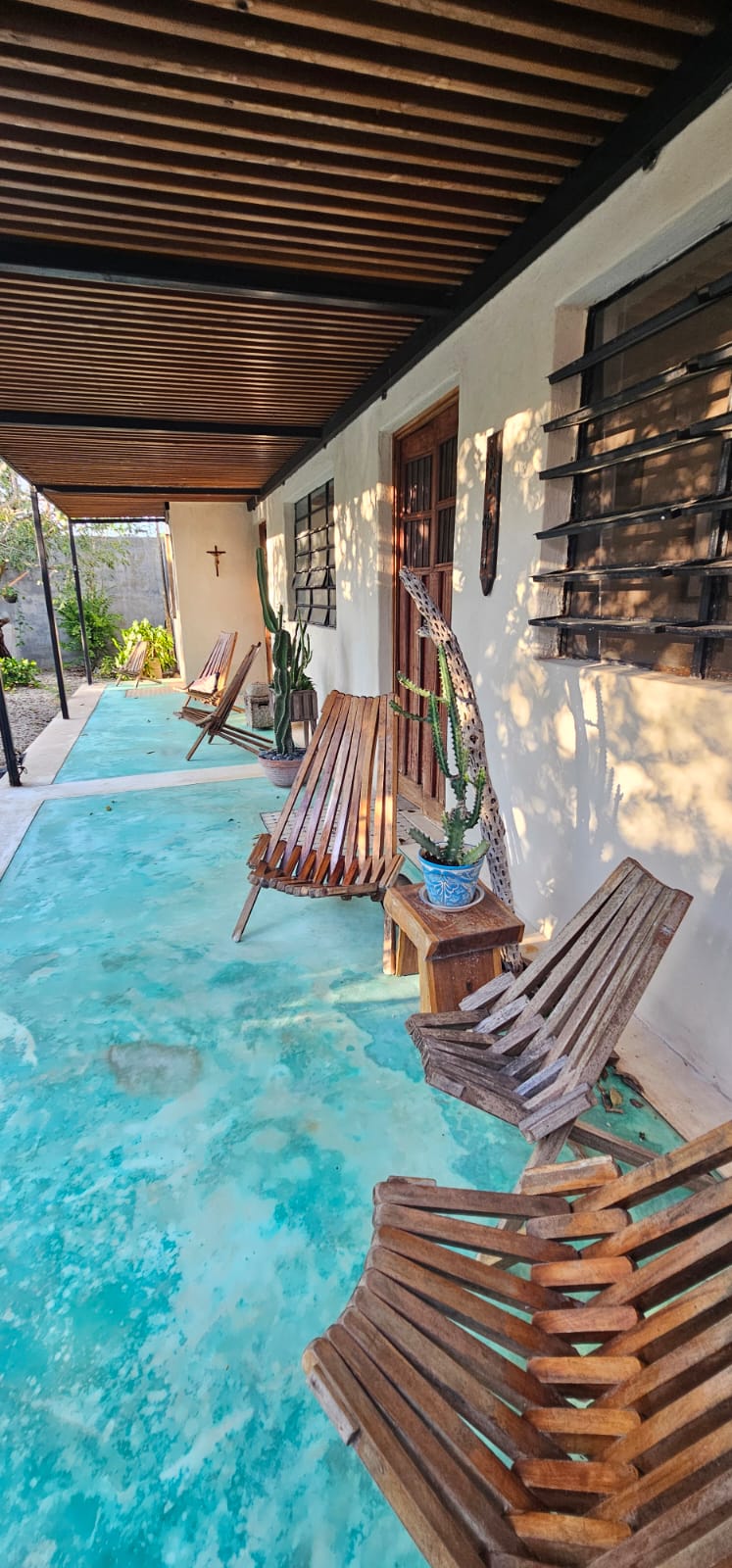 Patio exterior con piso turquesa, sillas de madera, cactus y techo de pérgola.