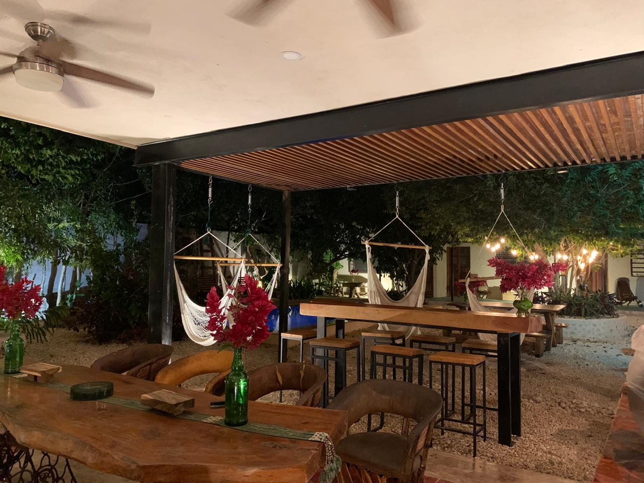 Patio exterior con mesas de madera, sillas, hamacas y luces decorativas por la noche.