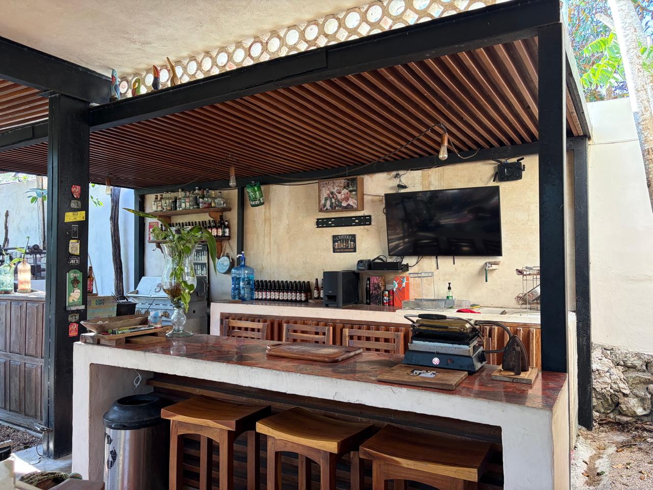 Cocina exterior con barra, taburetes de madera, estantes con botellas y televisión.