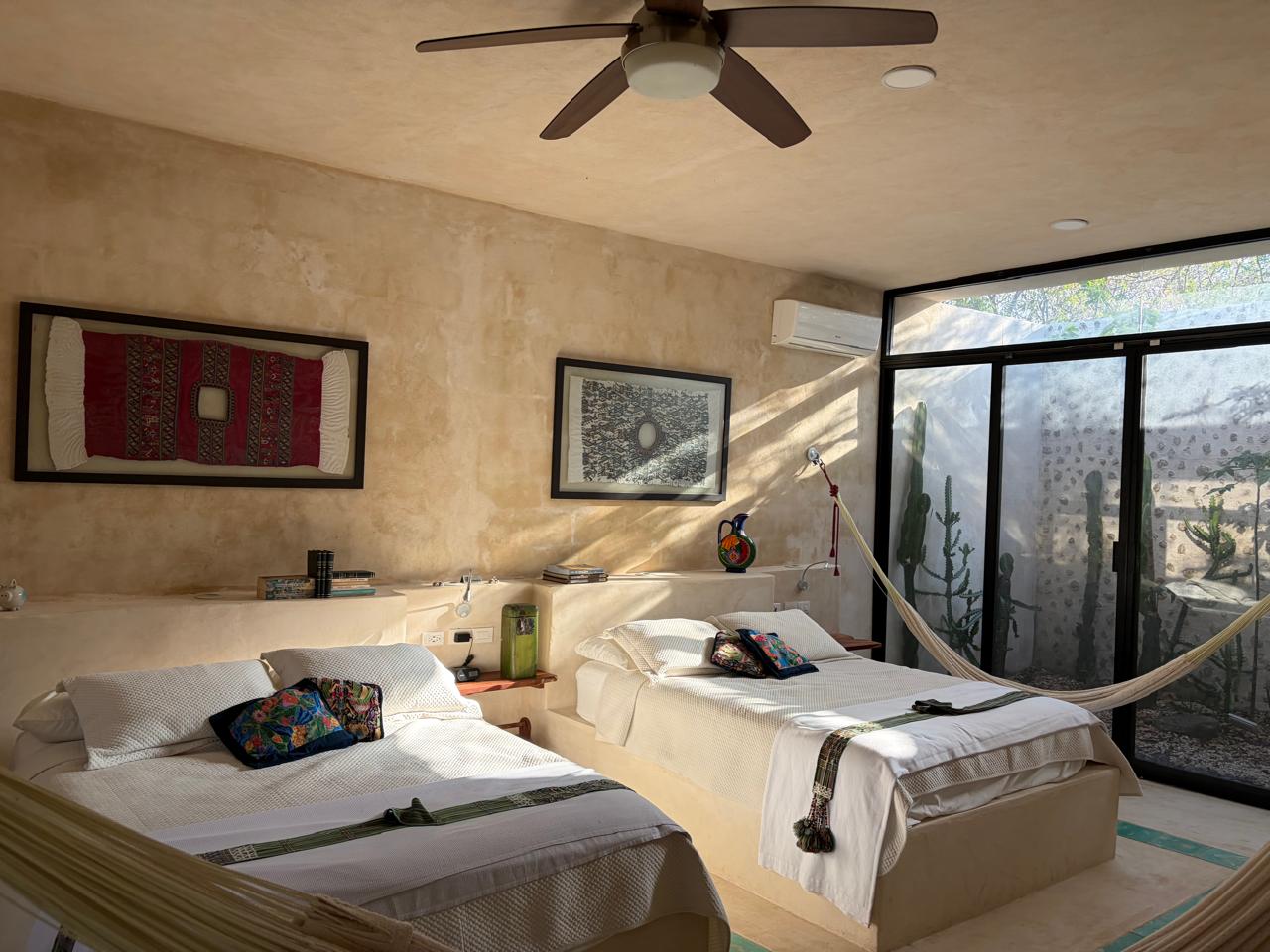 Recámara con dos camas individuales, hamacas, ventilador de techo y ventanal con vista a cactus.